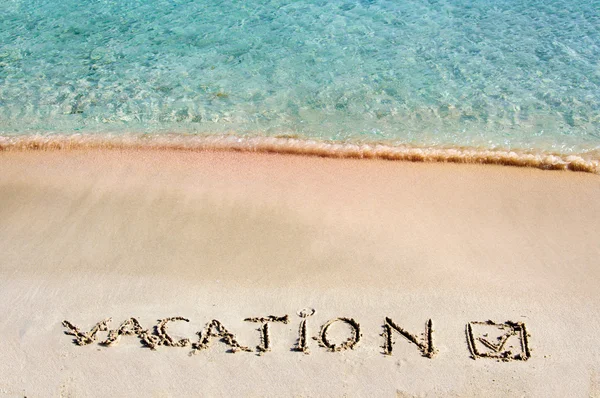 Vacation and checked mark written on sand on a beautiful beach, blue waves in background — Stock Photo, Image