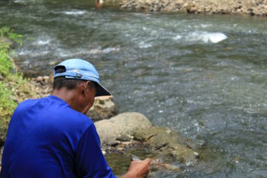 Bulanık nehir arka planında mavi gömlekli ve mavi şapkalı olgun Asyalı adamın dikiz görüntüsü