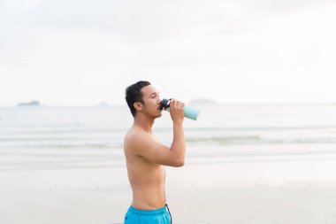 asian sport man drinking water while enjoying a morning run on the beach