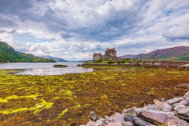 İskoç Dağları 'ndaki tarihi Eilean Donan Kalesi ile gölün kıyısı. Kaleye giden tarihi taş köprü. Gündüz vakti İskoçya 'da bulutlu gökyüzü olan eski bir kale