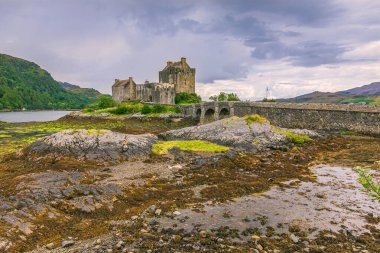 İskoçya 'da Eilean Donan Şatosu' nun tarihi binası. Gündüz vakti su alçak ve yayalar için taş bir köprü var. Dalgalar çekildiğinde ön planda yosun olan taşlar. Gökyüzündeki kara bulutlar