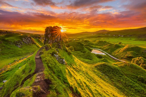 İskoçya 'daki Skye Adası' nda gün batımı. Dağ tepesinde güneş ışınları. Gökyüzünde kırmızı ve turuncu bulutlar. Ewen Şatosu 'nun geceleri patikaları ve çayırları var. Yolu olan küçük bir göl. Vadide gün ışığı