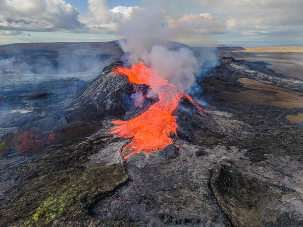 Red glowing lava flows from a volcano in Iceland's Reykjanes Peninsula. View of the crater from above. Steam and smoke near the crater. cooled magma around craters. clouds in the sky at day