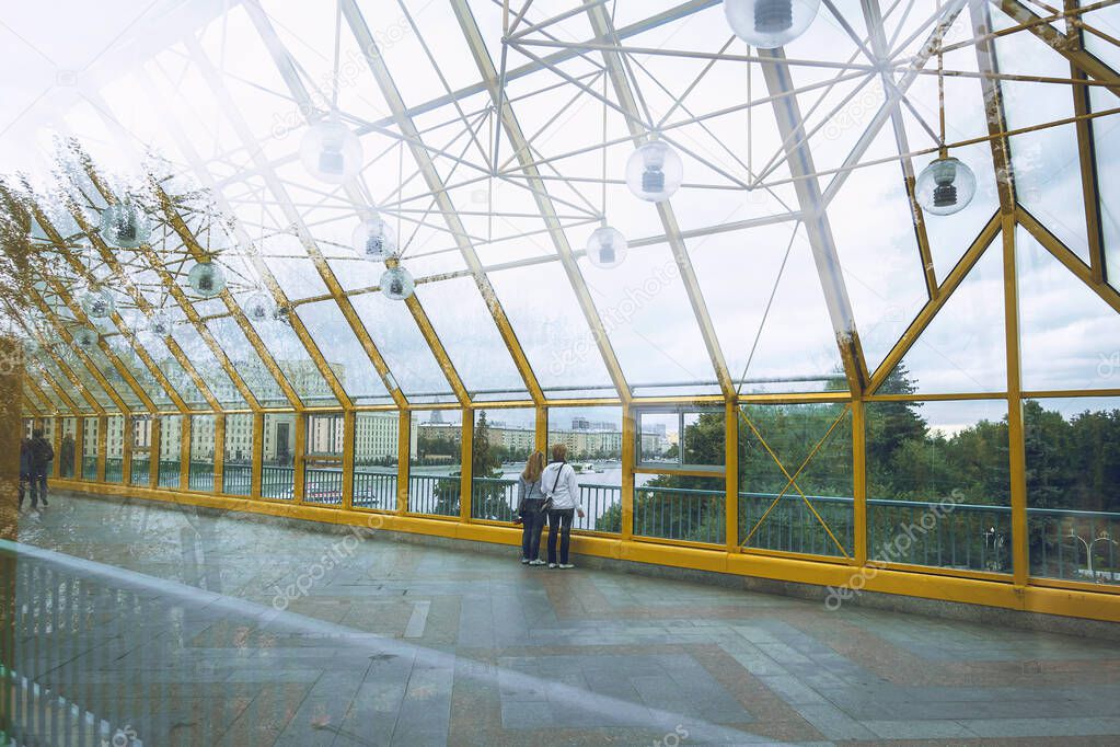 The fragment of glass pedestrian Andrew's bridge over the Moscow river with yellow metal structures and round lanterns. Pedestrians admire the view from the bridge.