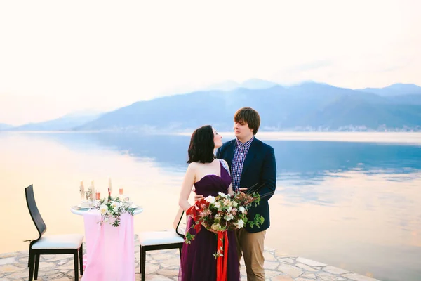 Man knuffelt vrouw met een boeket bloemen tegen de achtergrond van een serveertafel met stoelen op de pier tegen de achtergrond van water en bergen — Stockfoto