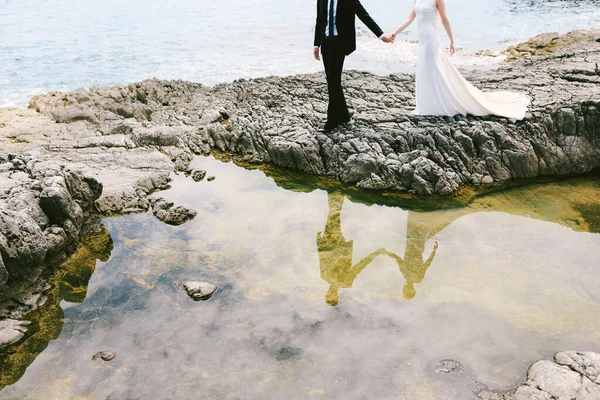 Novio y novia en un vestido blanco tomados de la mano, de pie en la orilla de un pequeño lago en una orilla rocosa cerca del mar —  Fotos de Stock