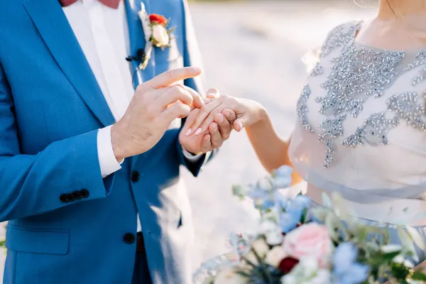 Lo sposo in un vestito blu mette una fede nuziale sul dito di spose con un mazzo di fiori. Primo piano — Foto Stock