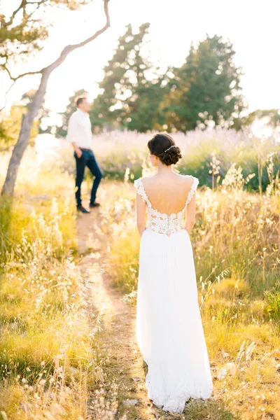 Sibenik, Kroatien - 05.06.17: Braut im weißen Spitzenkleid steht auf einem Weg in der Mitte des Feldes. Im Hintergrund steht Bräutigam mit den Händen in den Taschen. Rückseite — Stockfoto