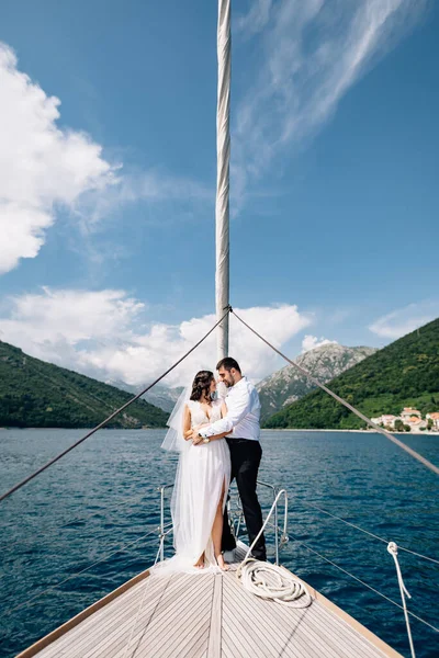 Nevěsta a ženich se objímají, zatímco stojí na přídi bílé jachty plachtění v zálivu Kotor — Stock fotografie