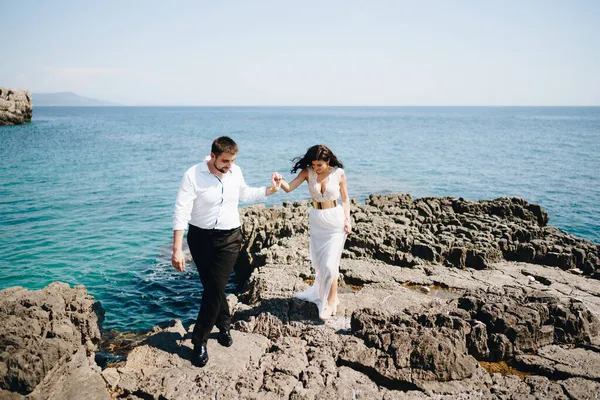 신랑 신부가 바위가 많은 해변에서 손을 잡고 걷고 있다 — 스톡 사진
