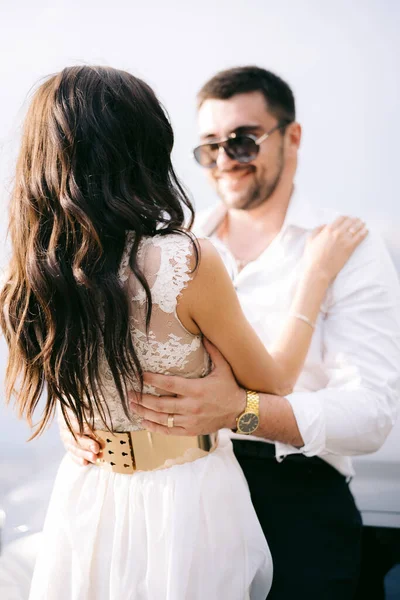 O noivo em óculos de sol gentilmente abraça a noiva e sorri. Mãos do noivo na cintura da noiva, close-up — Fotografia de Stock