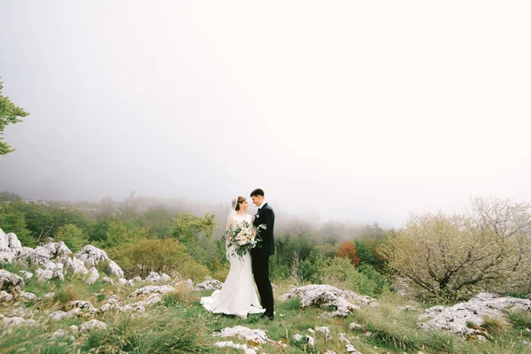 Bruid omhelst bruidegom in een lange jurk met een sluier en een boeket bloemen op een achtergrond van stenen en groen — Stockfoto