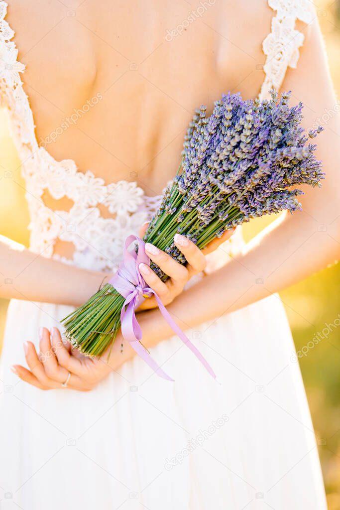 Bride in a white lace dress stands in a field and holds a lavender bouquet tied with a lilac ribbon behind her back. Close-up