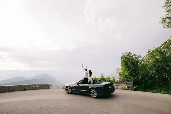 La novia y el novio están de pie con las manos en alto en un coche abierto en las pintorescas montañas de Montenegro — Foto de Stock