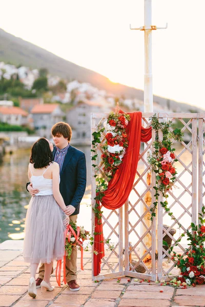 Bruid omhelst bruidegom met boeket bloemen op de pier in de buurt van een opengewerkte scherm versierd met bloemen en een rood gordijn tegen de achtergrond van water en bergen — Stockfoto