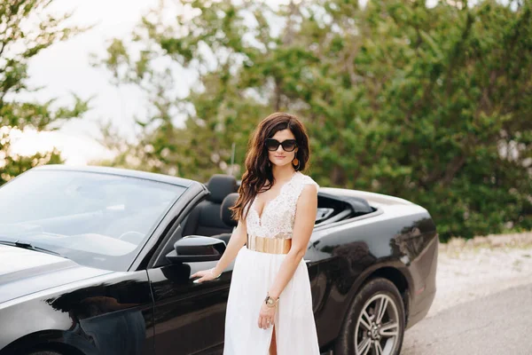 Стильная невеста в солнечных очках стоит рядом с черным кабриолетом на дороге в горах — стоковое фото