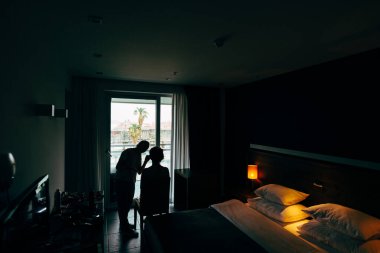 Makyaj sanatçısı yatak odasındaki pencerenin önünde müşteriyle barışır. Odada alacakaranlık