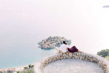 Kotor, Karadağ - 08.06.17: Burgundy elbiseli beyaz gömlekli damat gözlem güvertesinde oturup Sveti Stefan adasına bakıyor. Budva, Karadağ