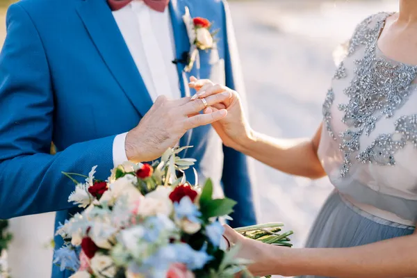 Bride in a lace dress puts an engagement ring on the finger of a groom in a blue suit. Close up — Stock Photo, Image