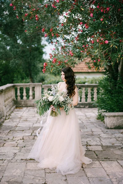 Bruden står under en blommande oleander med en bukett vita rosor, pioner, delfiner och eukalyptus grenar i händerna — Stockfoto