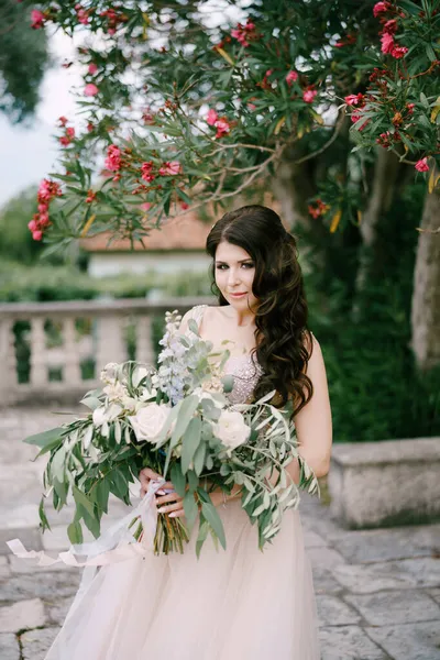 Pengantin wanita berdiri di bawah sebuah oleander mekar dengan buket mawar putih, peoni, delphinium dan cabang eukaliptus di tangannya — Stok Foto