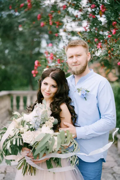 La mariée et le marié s'embrassent sous une laurier rose en fleurs, la mariée tient un bouquet dans ses mains — Photo