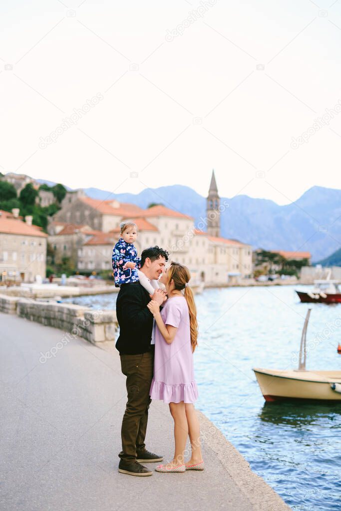 Mom almost kisses smiling dad with daughter on his shoulders on the coast against the background of the sea, boats and ancient buildings of the town of Perast