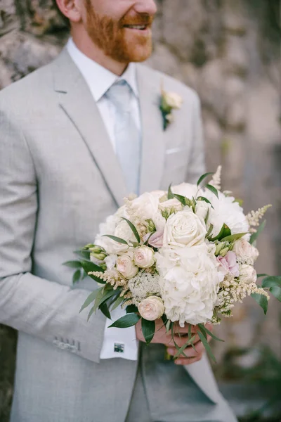 Groom avec un bouquet de fleurs se dresse contre un mur de pierre — Photo