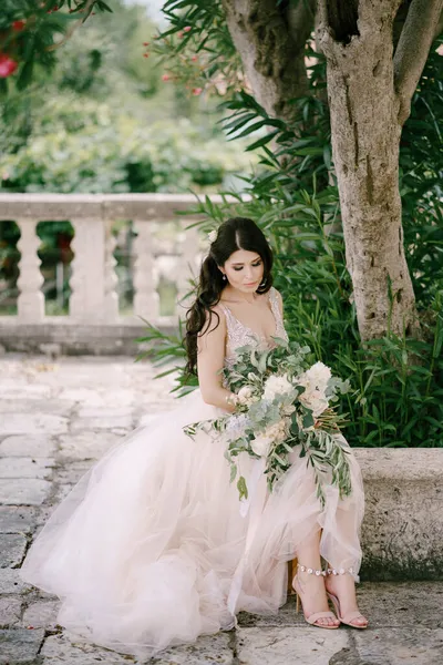 A noiva sentada perto do oleandro florescendo na cidade velha com um buquê de rosas brancas, peônias e galhos de eucalipto em suas mãos — Fotografia de Stock