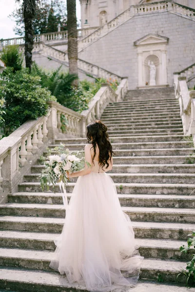 La novia está en las escaleras de la antigua iglesia en Prcanj con un ramo de novia en sus manos, vista trasera —  Fotos de Stock