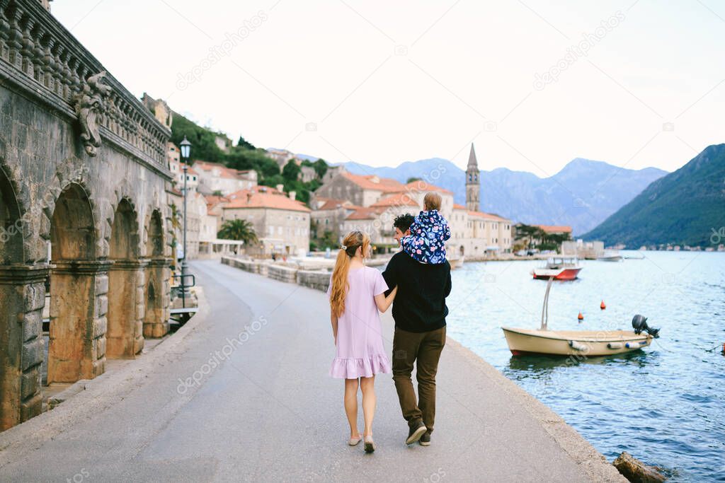 Mom and dad with a daughter on his shoulders walk along the coast against the background of the sea, mountains and houses of the town of Perast. Back view