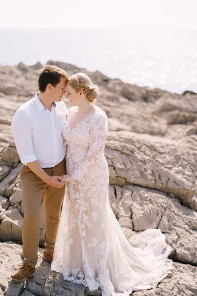 Abraço de noivo e noiva em pé na costa rochosa perto da água cintilante — Fotografia de Stock