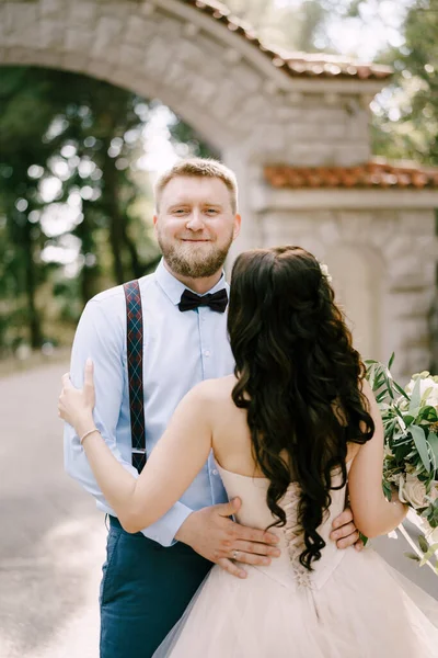 Nevěsta objímá ženicha v parku vedle krásného cihlového oblouku, pohled zezadu — Stock fotografie