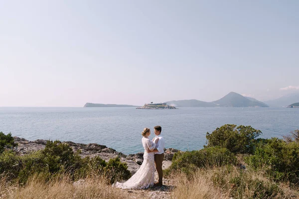 Esguicho e noiva estão no litoral contra o pano de fundo das montanhas e da ilha — Fotografia de Stock