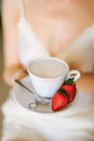 Chica en un vestido blanco sostiene en sus manos un platillo con una taza de capuchino, dos fresas, una cucharadita y un anillo. Primer plano — Foto de Stock