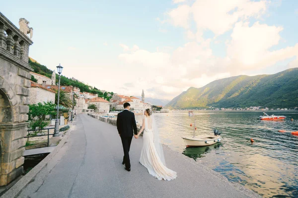 Mariée et marié marchent le long de la jetée en se tenant la main, sur fond de baie, de bateaux et de bâtiments anciens — Photo