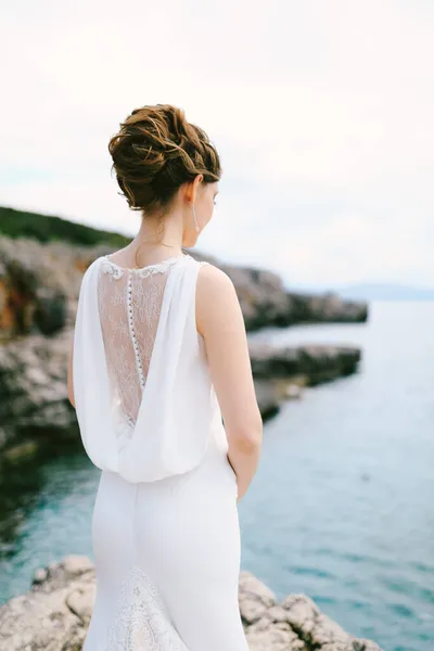 Pengantin wanita yang termenung dengan gaun putih yang berenda berdiri di pantai batu di atas laut. Tampilan belakang — Stok Foto