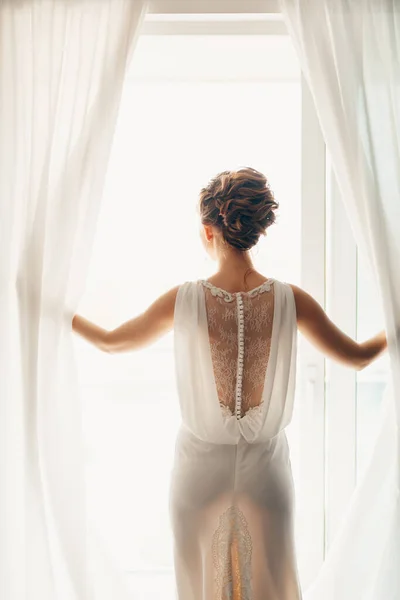 Noiva em um vestido de renda branca com um penteado alto bonito espalha as cortinas na janela com as mãos — Fotografia de Stock