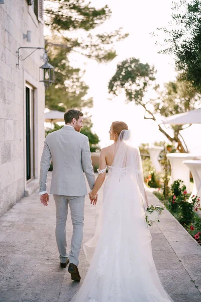 A menyasszony és a vőlegény kéz a kézben sétálnak az épület közelében a parkban — Stock Fotó