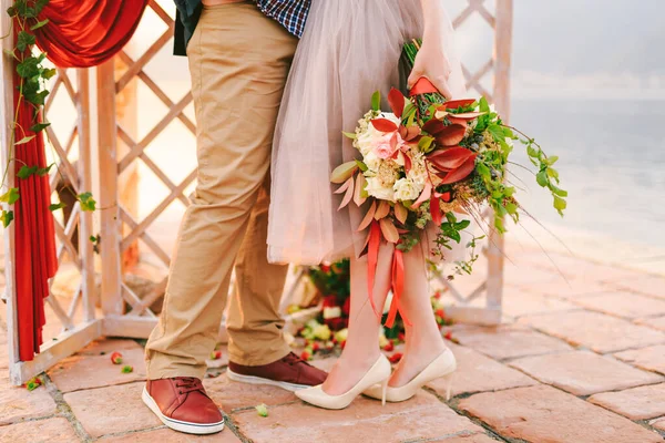 Bräutigam und Braut mit einem Blumenstrauß stehen auf einer Steinfliese vor dem Hintergrund einer geschnitzten Leinwand, die mit rotem Vorhang und Grün dekoriert ist. Nahaufnahme — Stockfoto