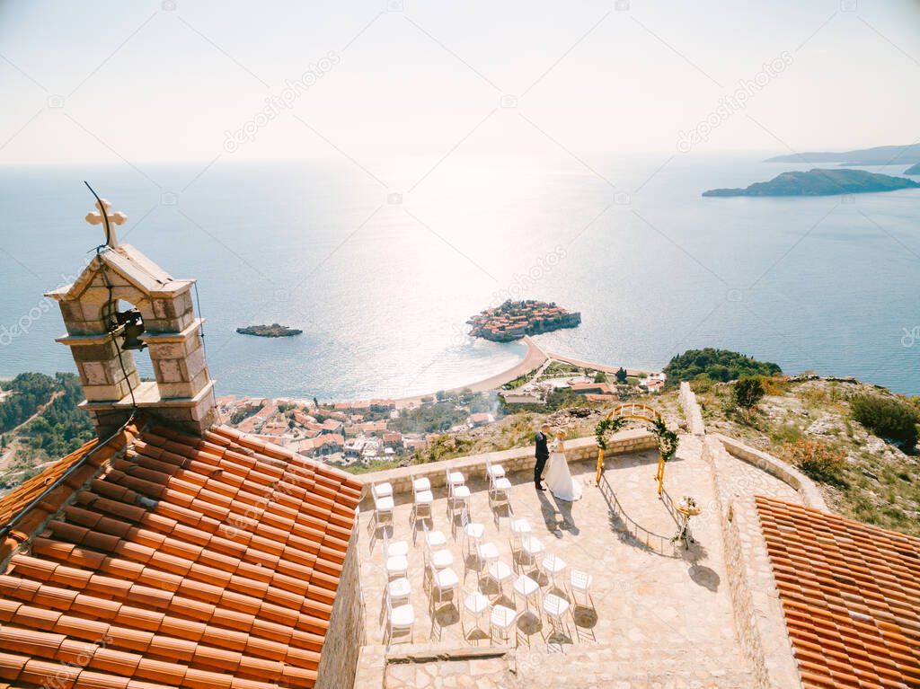 Bride and groom stand on the observation deck in front of the Sveti Sava church overlooking the Sveti Stefan island. Montenegro. View from above
