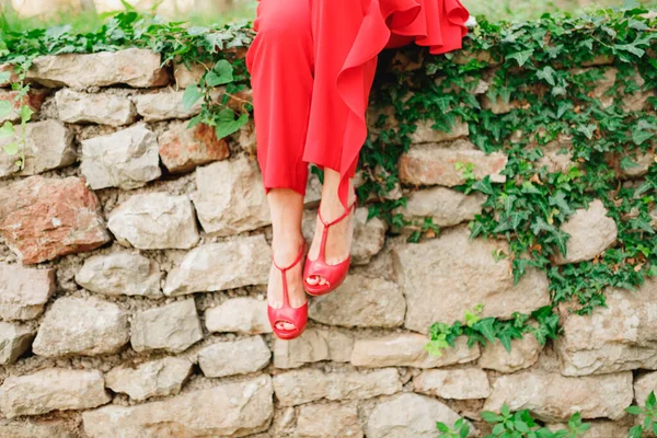 Vrouw in rode broek en rode sandalen zit op een stenen hek verstrengeld met klimop. Close-up — Stockfoto