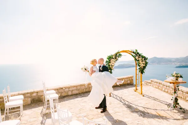 A vőlegény fehér ruhában tartja a menyasszonyt egy csokor virággal a karjában, miközben a kilátóteraszon áll, kilátással a Sveti Stefan szigetre. — Stock Fotó