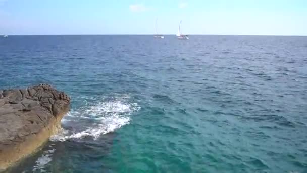 Волны бьют по скалистому берегу на фоне парусных судов — стоковое видео