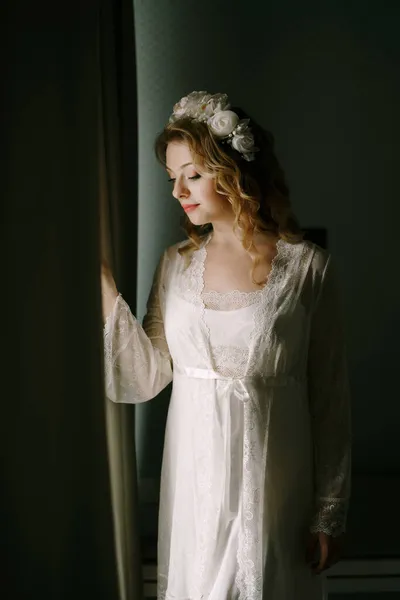 A bride in a white wreath and lace peignoir stands at the window and peeps out from behind the curtain in a hotel room — Stock Photo, Image