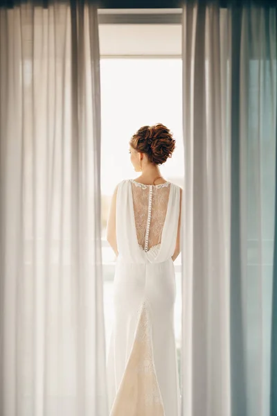 Noiva em um vestido de renda branca com um belo penteado alto fica na varanda. Vista do quarto — Fotografia de Stock