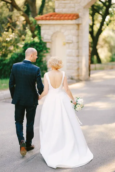 Γαμπρός σε μαύρο κοστούμι και νύφη σε λευκό φόρεμα με μπουκέτο λουλούδια περπατούν κρατώντας τα χέρια κατά μήκος του δρόμου προς την πέτρινη πύλη στον κήπο. Πίσω όψη — Φωτογραφία Αρχείου