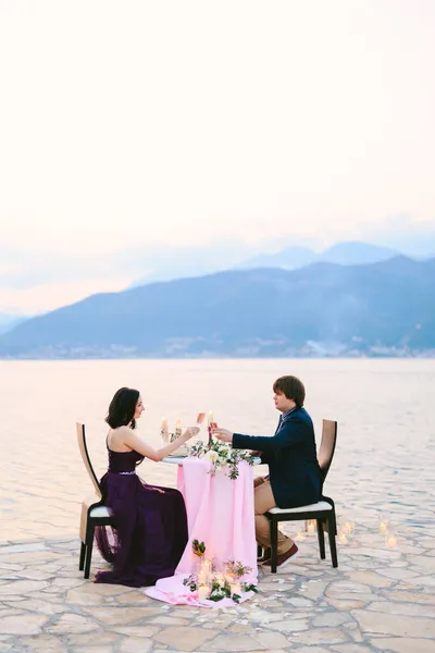 Man en vrouw zitten aan een tafel en knikken glazen op de pier tegen de achtergrond van de zee en de bergen — Stockfoto