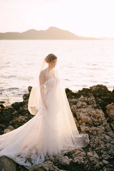 Sposa in abito bianco e veli si erge sulle rocce in riva al mare — Foto Stock