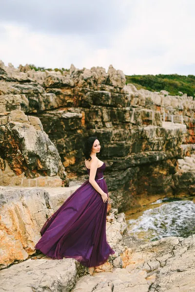 Rüzgarda çırpınan leylak elbiseli kız kaynayan suyun arka planında bir kayanın üzerinde duruyor. — Stok fotoğraf